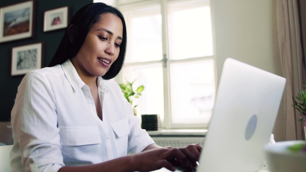 A young asian woman with laptop sitting at the desk indoors at home, typing.