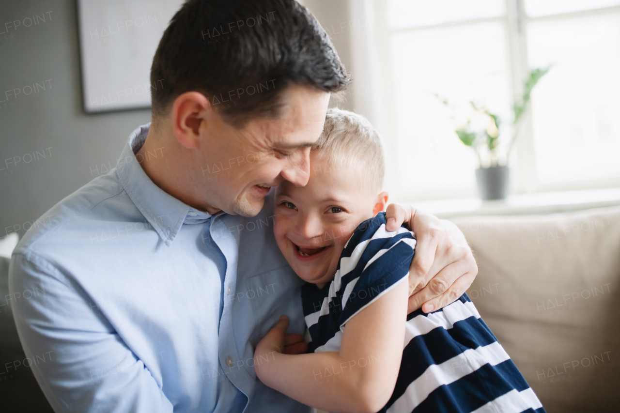 A father with happy down syndrome son indoors at home, hugging and laughing.