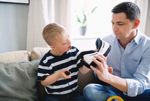 A father with happy down syndrome son indoors at home, using vr goggles.