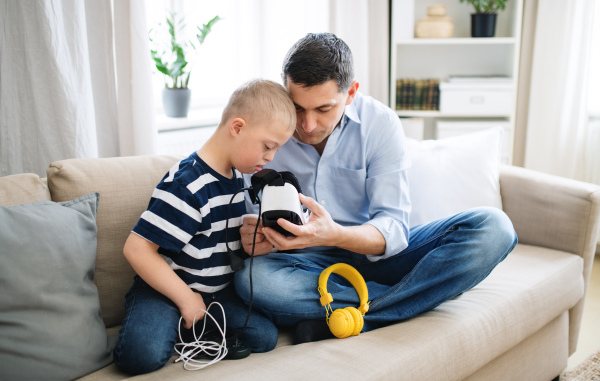 A father with happy down syndrome son indoors at home, using vr goggles.