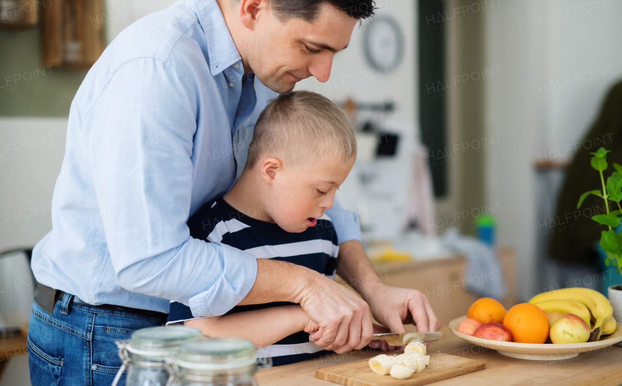 A father with happy down syndrome son indoors in kitchen, chopping fruit.