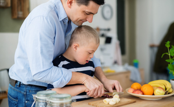 A father with happy down syndrome son indoors in kitchen, chopping fruit.