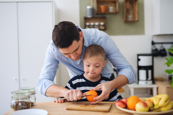 A father with happy down syndrome son indoors in kitchen, preparing food.