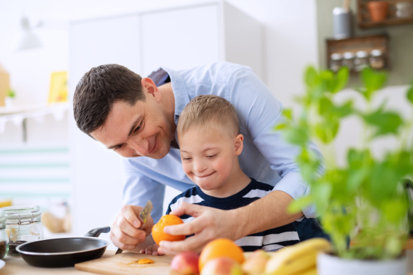 A father with happy down syndrome son indoors in kitchen, preparing food.