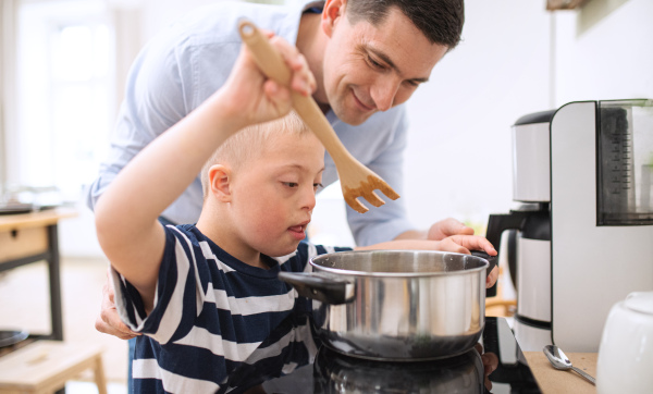 A father with happy down syndrome son indoors in kitchen, cooking.