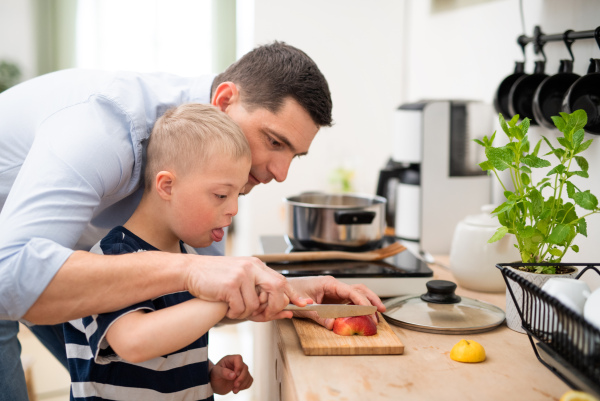 A father with happy down syndrome son indoors in kitchen, chopping apple.