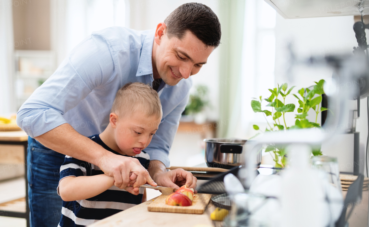 A father with happy down syndrome son indoors in kitchen, chopping apple.