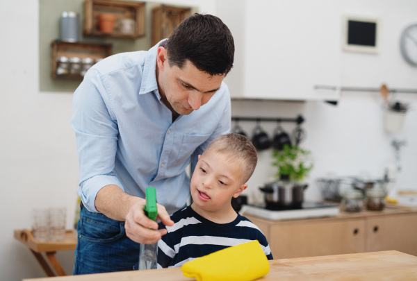 A father with happy down syndrome son indoors in kitchen, cleaning table.