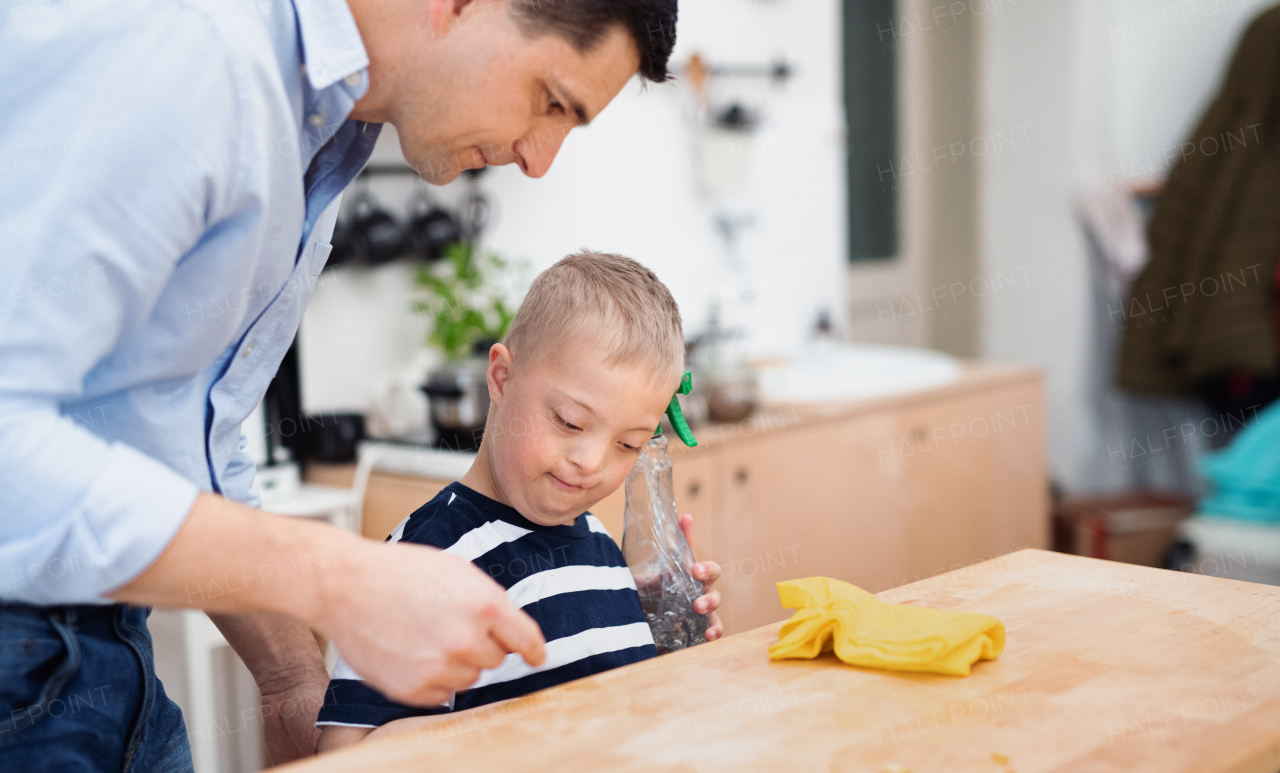 A father with happy down syndrome son indoors in kitchen, cleaning table.