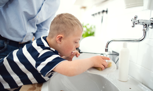 Am unrecognizable father with down syndrome son indoors in kitchen, washing dishes.