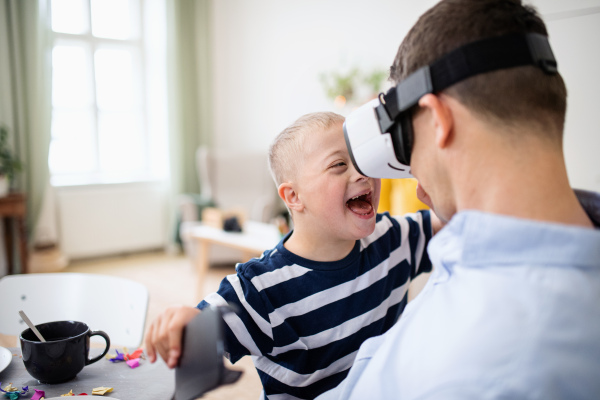 A father with happy down syndrome son indoors at home, using vr goggles.