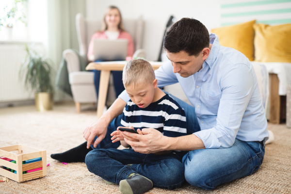 A father with happy down syndrome son indoors at home, using smartphone.