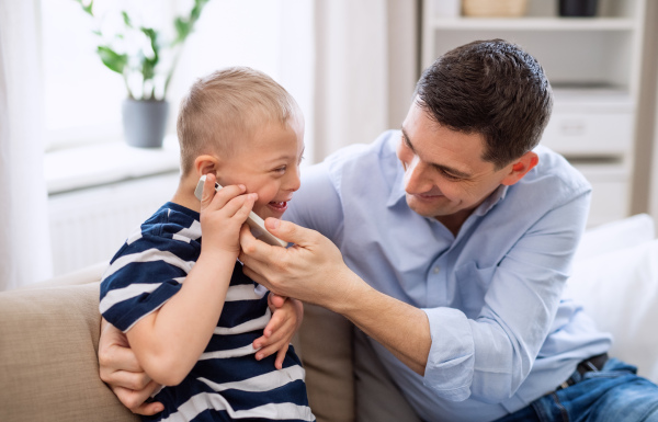 A father with happy down syndrome son indoors at home, using smartphone.