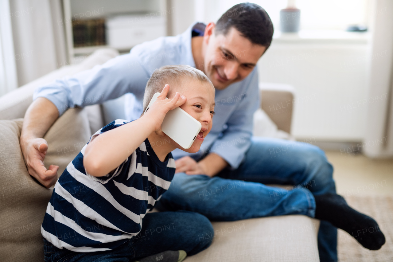 A father with happy down syndrome son indoors at home, using smartphone.