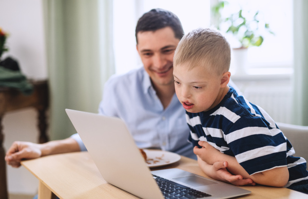 A father with happy down syndrome son indoors at home, using laptop.