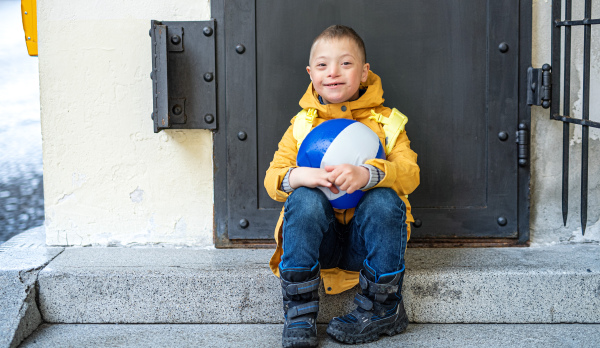 A happy down syndrome boy with ball outdoors sitting in front of door, looking at camera.