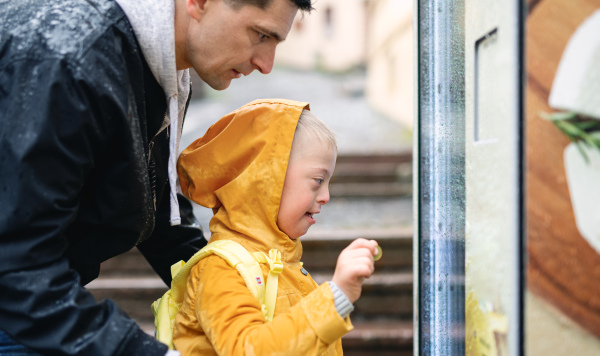 A happy down syndrome boy with father outdoors on a walk in rain, playing.