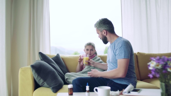 A mature father giving juice to his sick small daughter indoors at home.