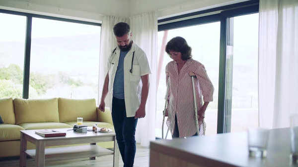 Health visitor and a senior woman with crutches during home visit, walking. Slow motion.