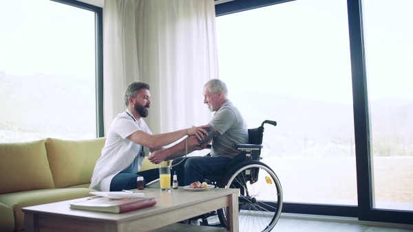 Health visitor and a senior man in wheelchair during home visit, checking blood pressure. Slow motion.