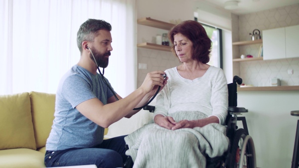 Health visitor examining a senior woman in wheelchair with stethoscope during home visit.