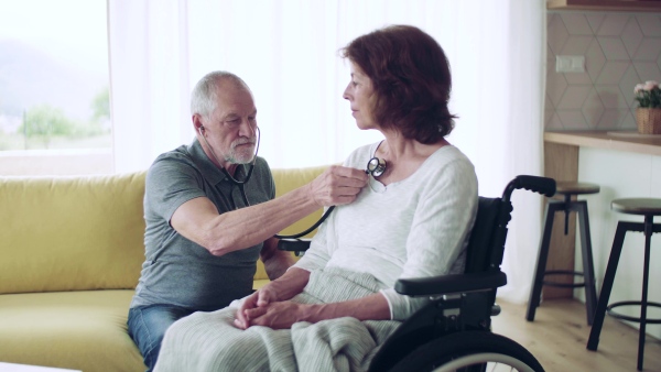 Health visitor examining a senior woman in wheelchair with stethoscope during home visit.