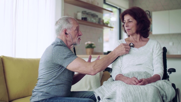 Health visitor examining a senior woman in wheelchair with stethoscope during home visit.