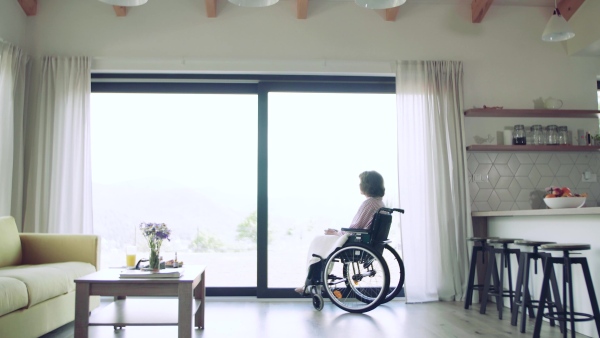 A lonely senior woman with wheelchair indoors at home looking out of window.