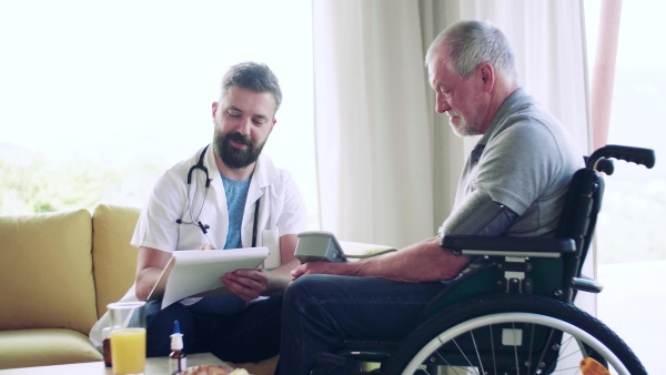 Health visitor and a senior man in wheelchair during home visit, checking blood pressure.