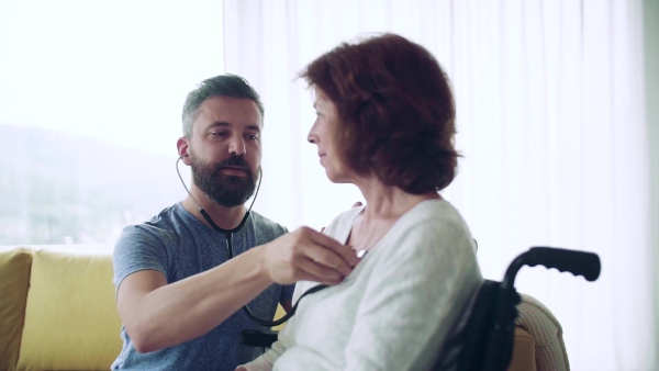 Health visitor examining a senior woman in wheelchair with stethoscope during home visit. Slow motion.