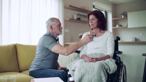 Health visitor examining a senior woman in wheelchair with stethoscope during home visit. Slow motion.