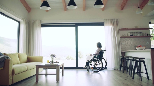 A lonely senior woman with wheelchair indoors at home looking out of window. Slow motion.