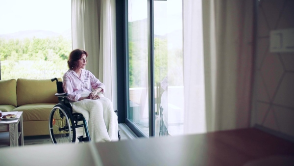 A lonely senior woman with wheelchair indoors at home looking out of window. Slow motion.