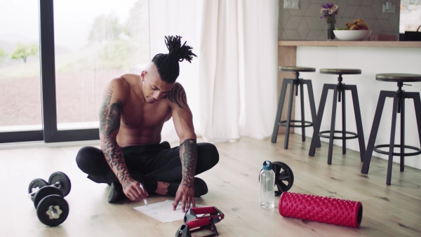 A mixed race young man with tattoo doing exercise indoors at home, keeping records.