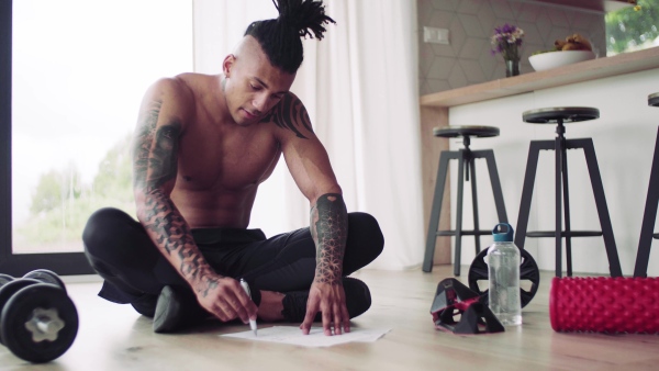 A mixed race young man with tattoo doing exercise indoors at home, keeping records.