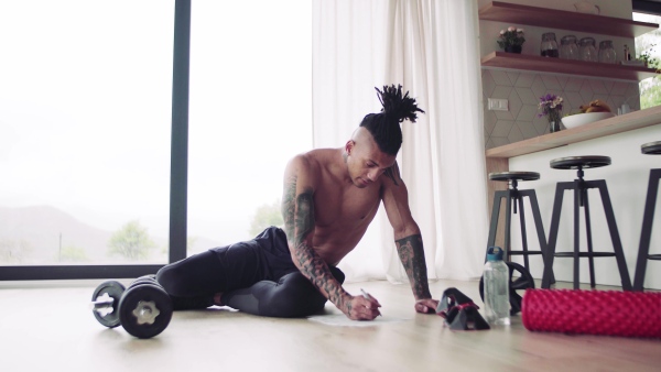 A mixed race young man with tattoo doing exercise indoors at home, keeping records.