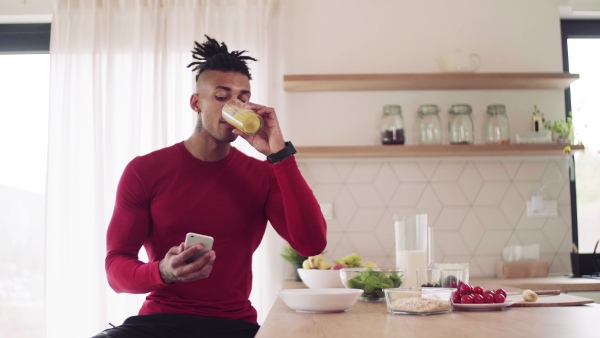 A mixed race young sporty man with smartphone indoors at home, drinking juice.