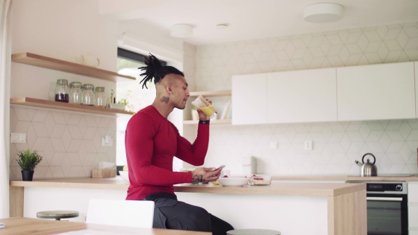 A mixed race young sporty man with smartphone indoors at home, drinking juice.