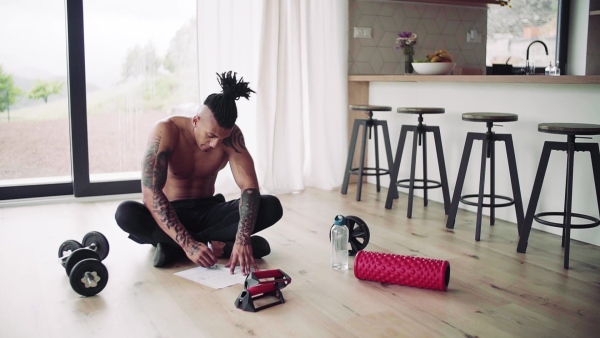 A mixed race young man with tattoo doing exercise indoors at home, keeping records. Slow motion.