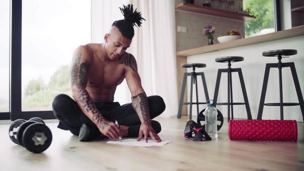 A mixed race young man with tattoo doing exercise indoors at home, keeping records. Slow motion.