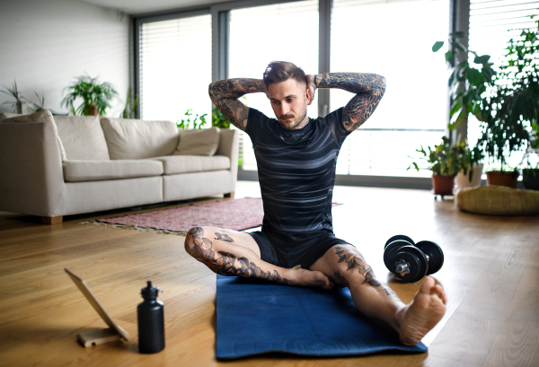 Front view portrait of young man with tablet doing workout exercise indoors at home.