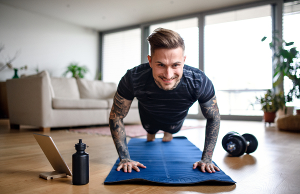 Front view portrait of young man with tablet doing workout exercise indoors at home.