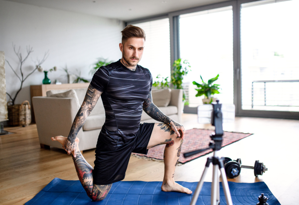 Young man trainer doing online workout exercise indoors at home, using camera.