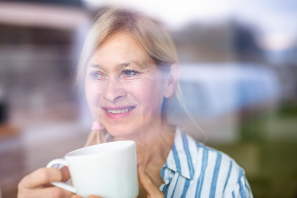 Portrait of senior woman with cup of coffee indoors at home. Shot through glass.