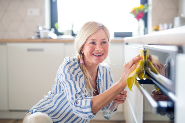 Portrait of happy senior woman cleaning oven door indoors in kitchen at home.