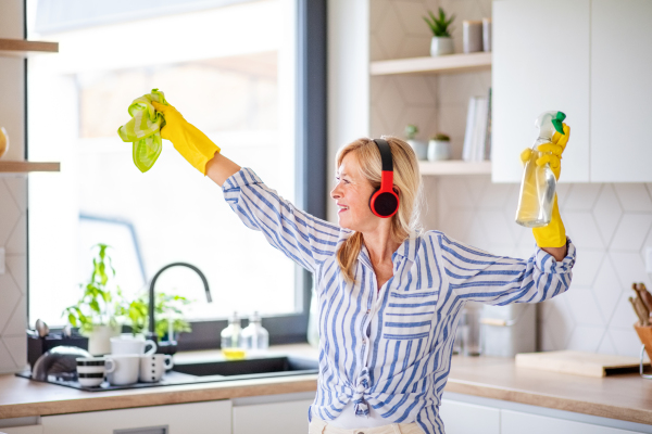 Portrait of cheerful senior woman with headphones and gloves cleaning indoors at home.