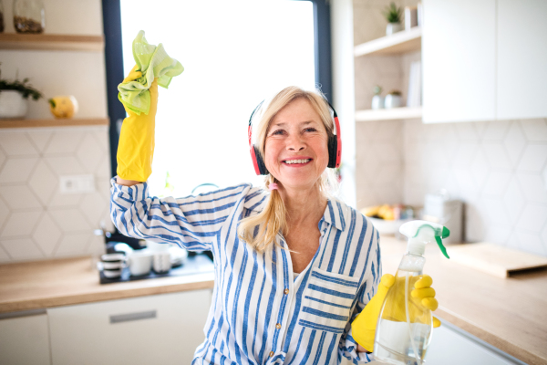 Portrait of cheerful senior woman with headphones and gloves cleaning indoors at home.