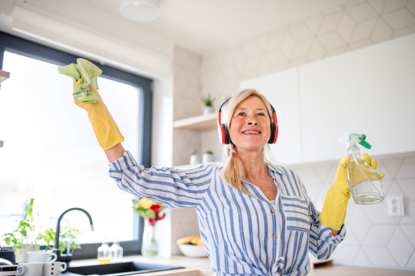 Portrait of cheerful senior woman with headphones and gloves cleaning indoors at home.