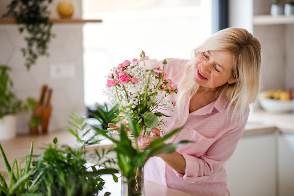 Portrait of happy senior woman arranging flowers in vase indoors at home.