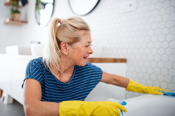 Happy senior woman with gloves cleaning bathroom indoors at home.